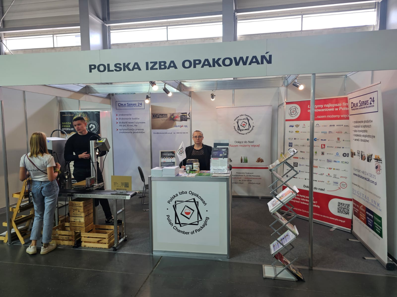 Stoisko targowe Polskiej Izby Opakowań z prezentacją drukarki do znakowania opakowań, mężczyzna demonstruje urządzenie klientce, w tle regał z ulotkami i materiałami reklamowymi.