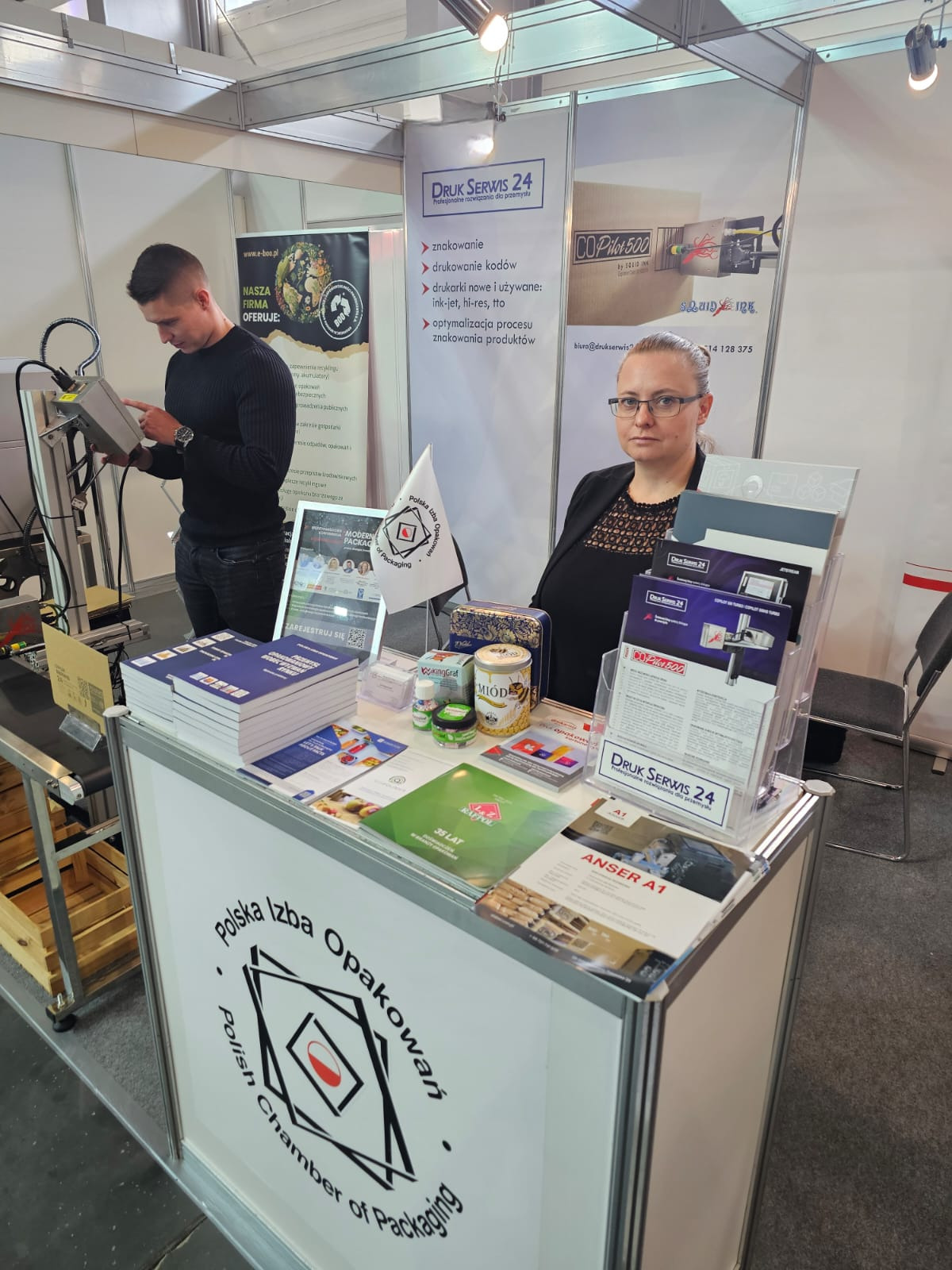 Stoisko targowe firmy Druk Serwis 24 z ekspozycją materiałów reklamowych, próbek i urządzeń do znakowania produktów, obsługiwane przez dwie osoby.