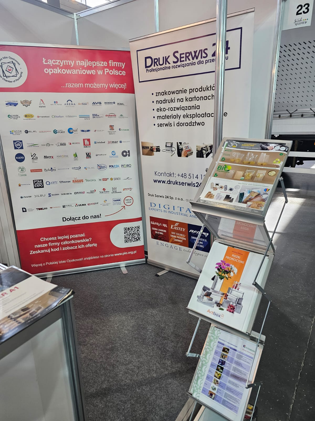 Stoisko targowe firmy oferującej usługi druku i znakowania opakowań, prezentacja materiałów reklamowych i katalogów produktowych, widoczne roll-upy z logo firmy i hasłami reklamowymi.
