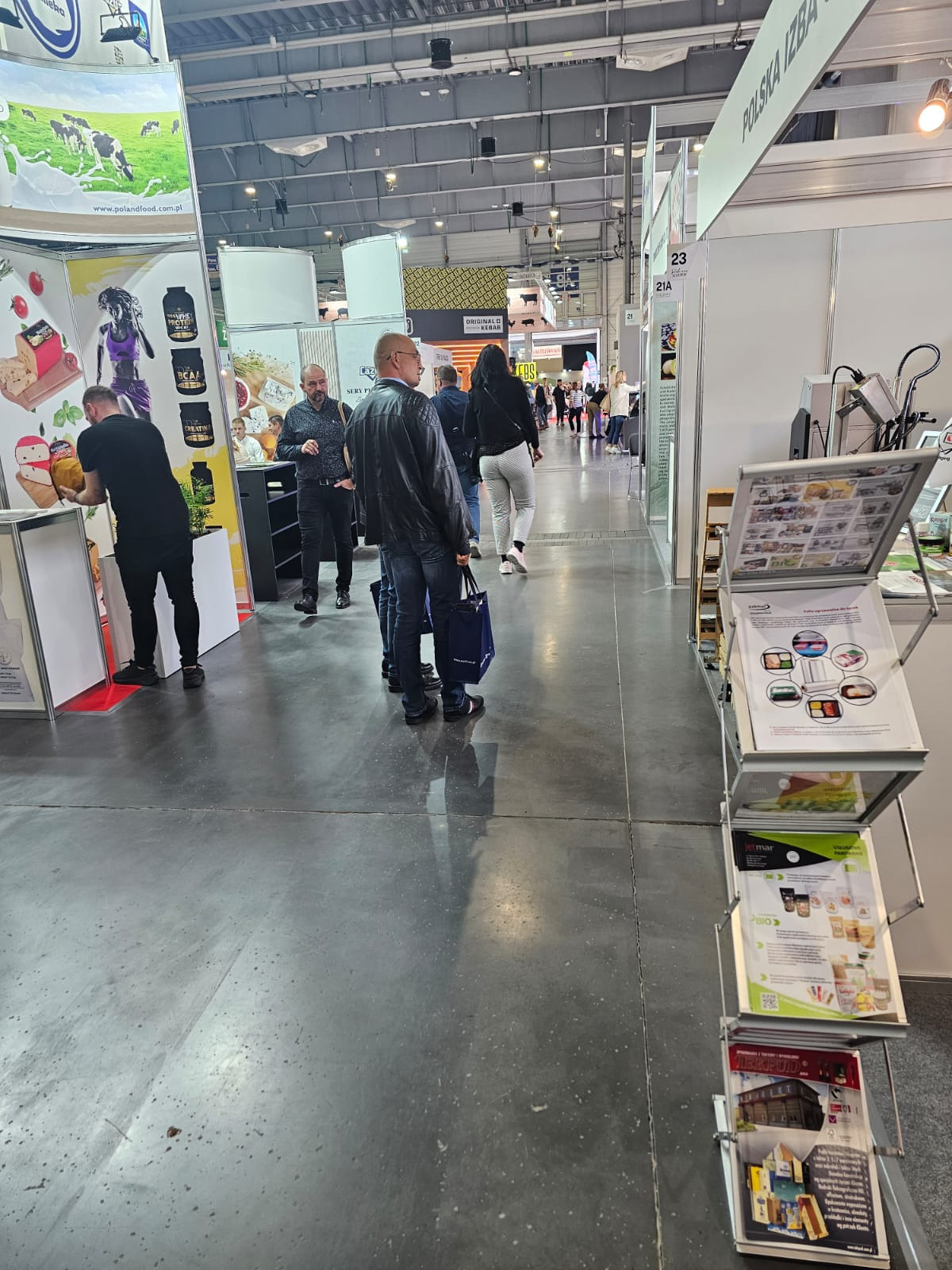 Wnętrze hali wystawienniczej podczas targów, widoczne stoiska firm z produktami spożywczymi, przechodzący zwiedzający i metalowy stojak z ulotkami reklamowymi w pierwszym planie.