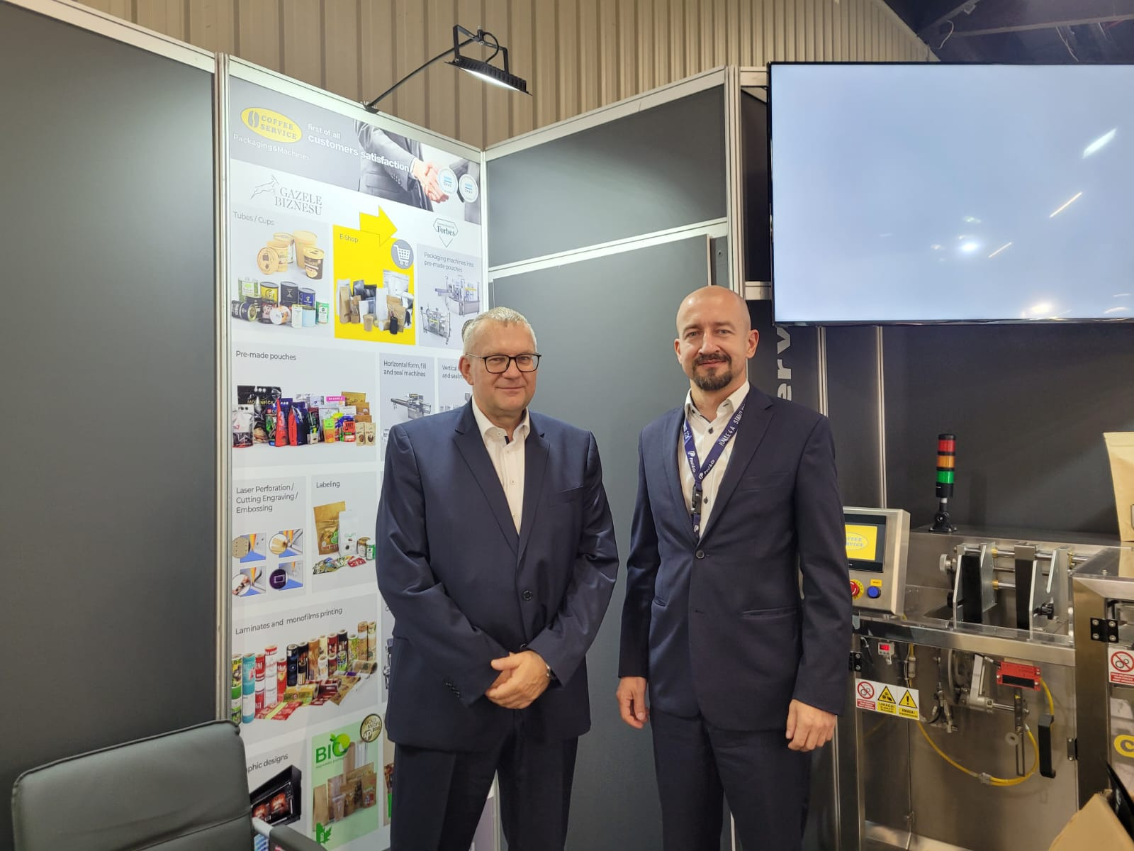 Dwóch mężczyzn w garniturach na stoisku targowym prezentującym maszyny do pakowania i przykłady opakowań produktów spożywczych, z widoczną maszyną pakującą w tle.