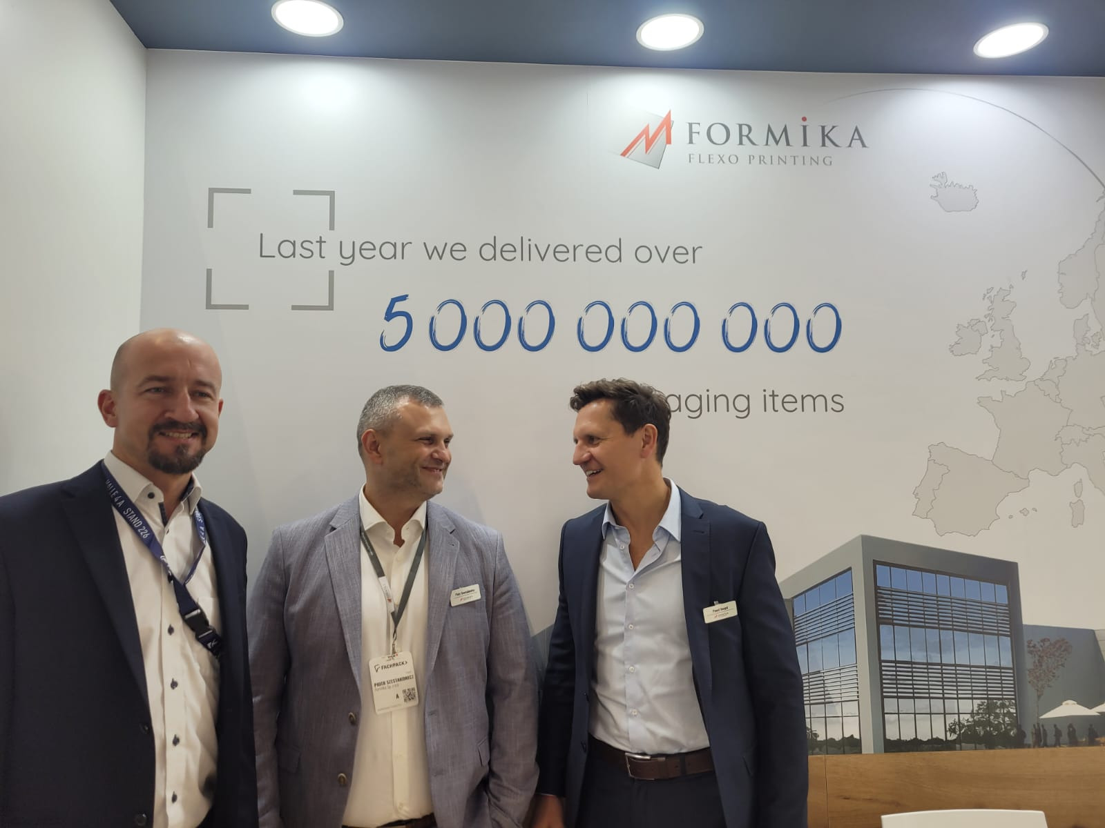 Trzech mężczyzn w garniturach uśmiecha się do siebie na tle ściany z logo firmy Formika Flexo Printing i informacją o dostarczeniu ponad 5 miliardów opakowań, widoczna mapa Europy i grafika budynku.