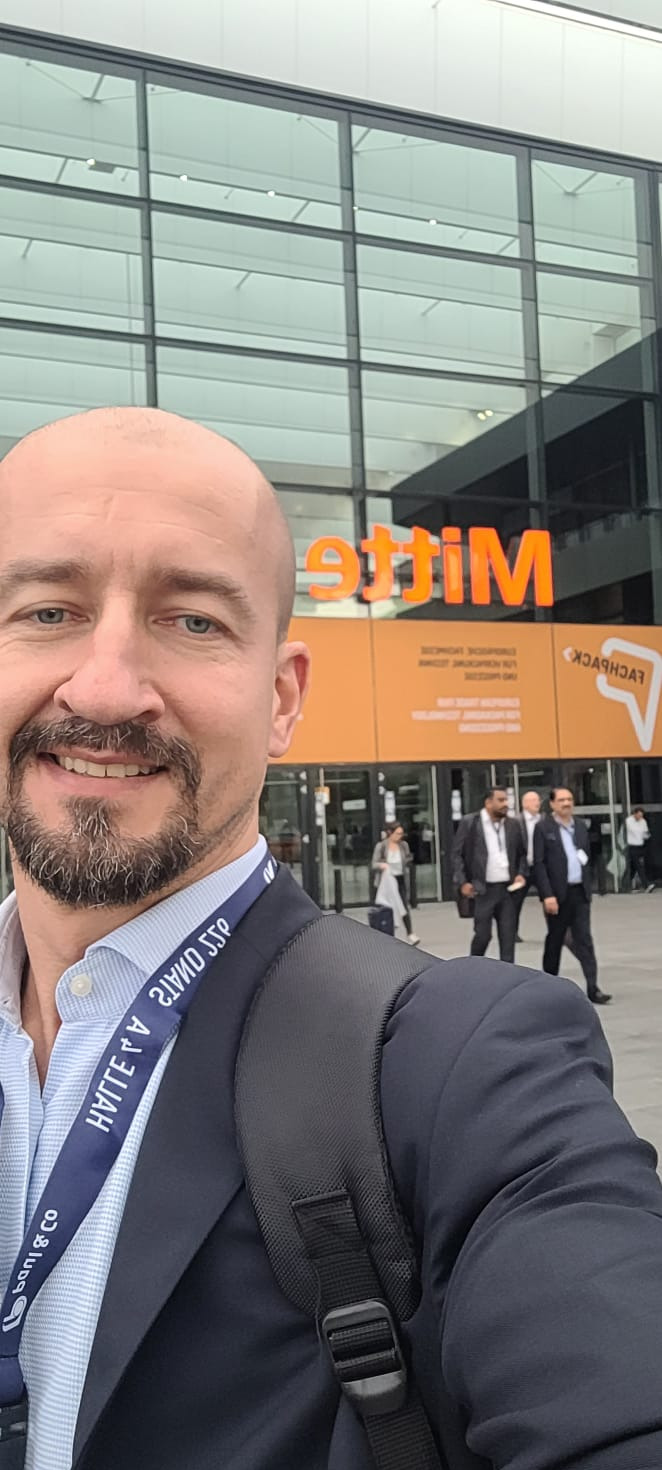 Portret mężczyzny na tle wejścia do hali targowej w Warszawie, z widocznym napisem 'Mitte' i logo 'Fachpack', sugerujący udział w wydarzeniu branżowym związanym z pakowaniem.