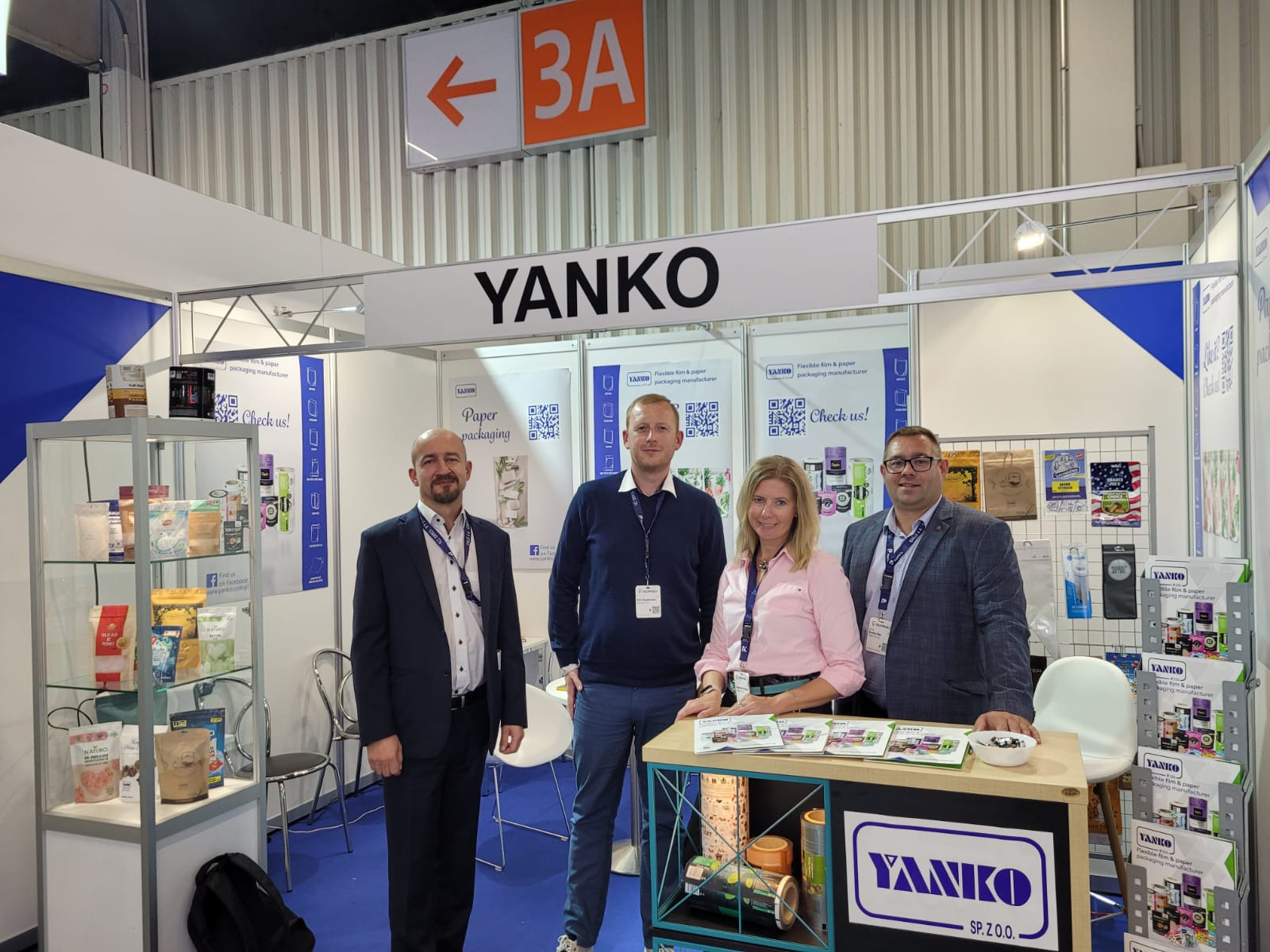 Stoisko targowe firmy Yanko z pracownikami prezentujące opakowania papierowe i foliowe na produkty spożywcze, widoczne ekspozytory z produktami, ulotki i logo firmy.