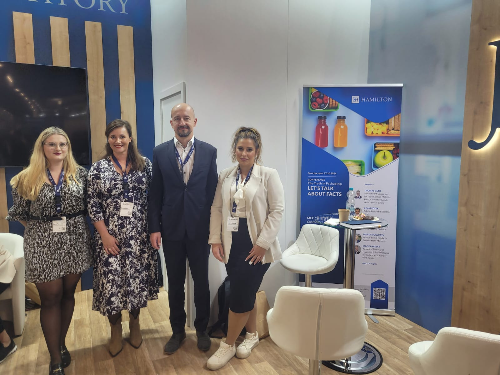 Grupa osób na stoisku targowym z banerem reklamującym konferencję o opakowaniach, prezentującym zdjęcia zapakowanych produktów spożywczych.