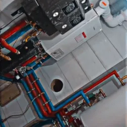 Kompleksowa instalacja kotła grzewczego z licznymi rurami w kolorach niebieskim i czerwonym, widok z dołu na tle białych płytek ceramicznych.