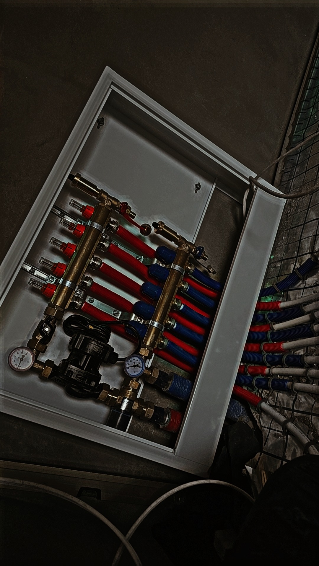 Rozdzielacz ogrzewania podłogowego w metalowej szafce z widocznymi rurami w kolorze czerwonym i niebieskim, manometry wskazujące ciśnienie, instalacja w trakcie montażu.