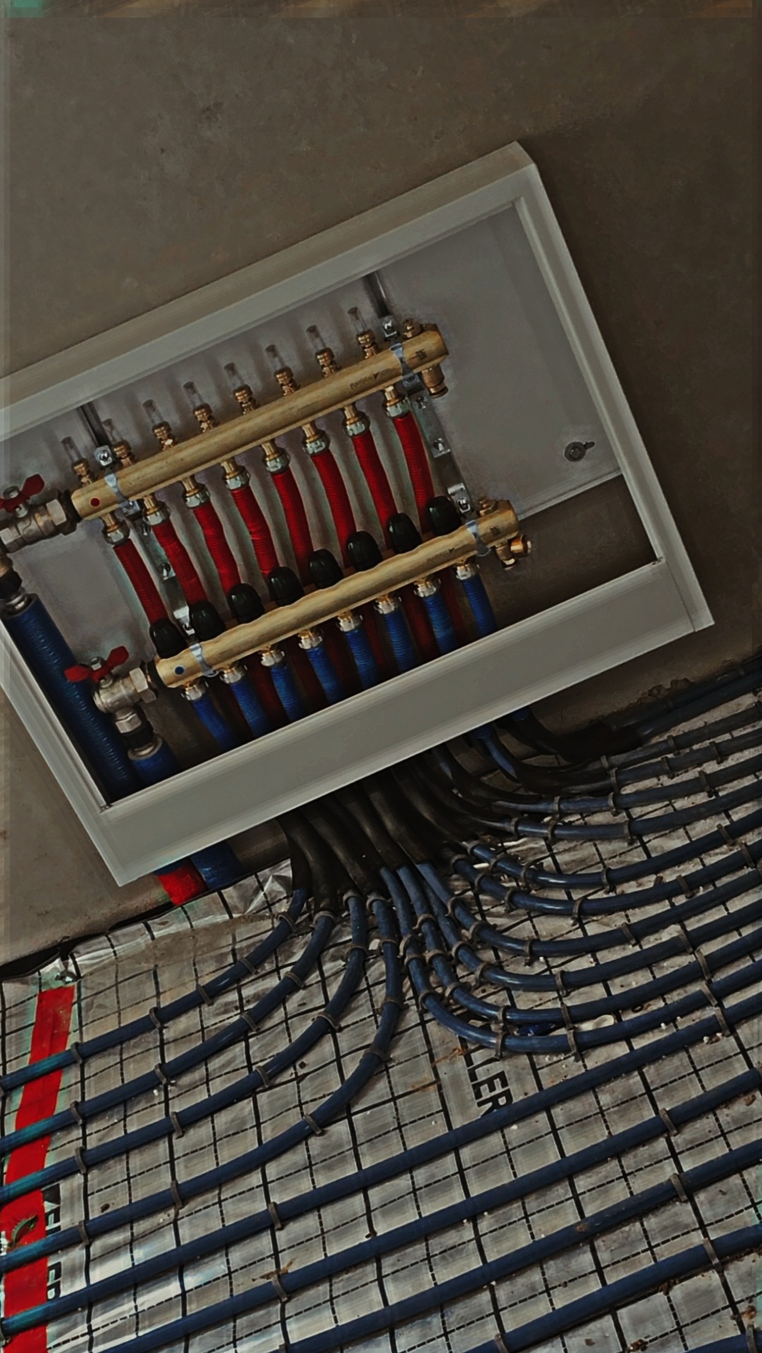 Rozdzielacz z rurami czerwonymi i niebieskimi w skrzynce, wychodzące rury do ogrzewania podłogowego na siatce montażowej, widok z góry pod kątem.