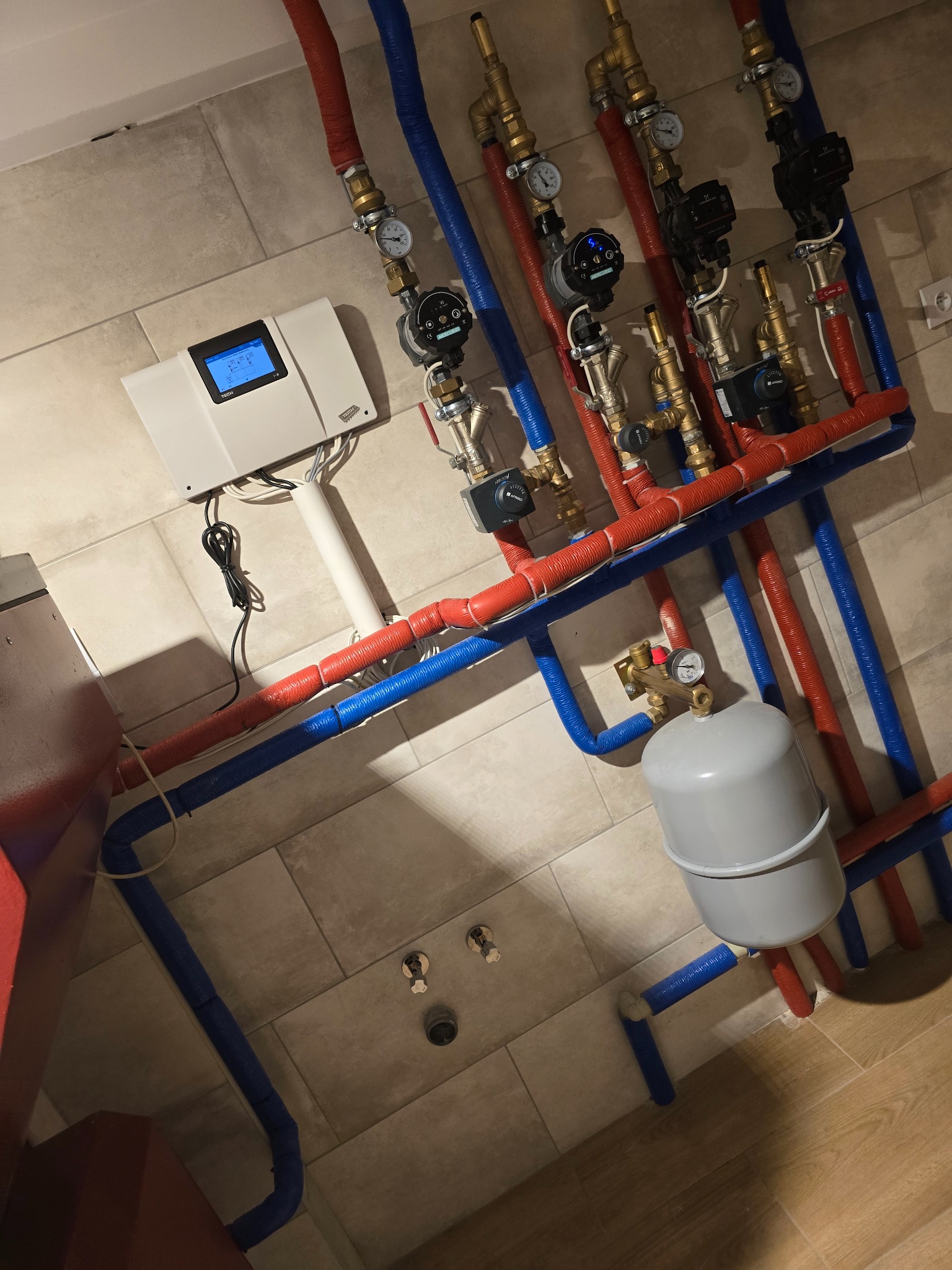 Rozbudowana instalacja centralnego ogrzewania z czerwono-niebieskimi rurami, pompami obiegowymi, zbiornikiem wyrównawczym i elektronicznym sterownikiem na tle płytek ceramicznych.