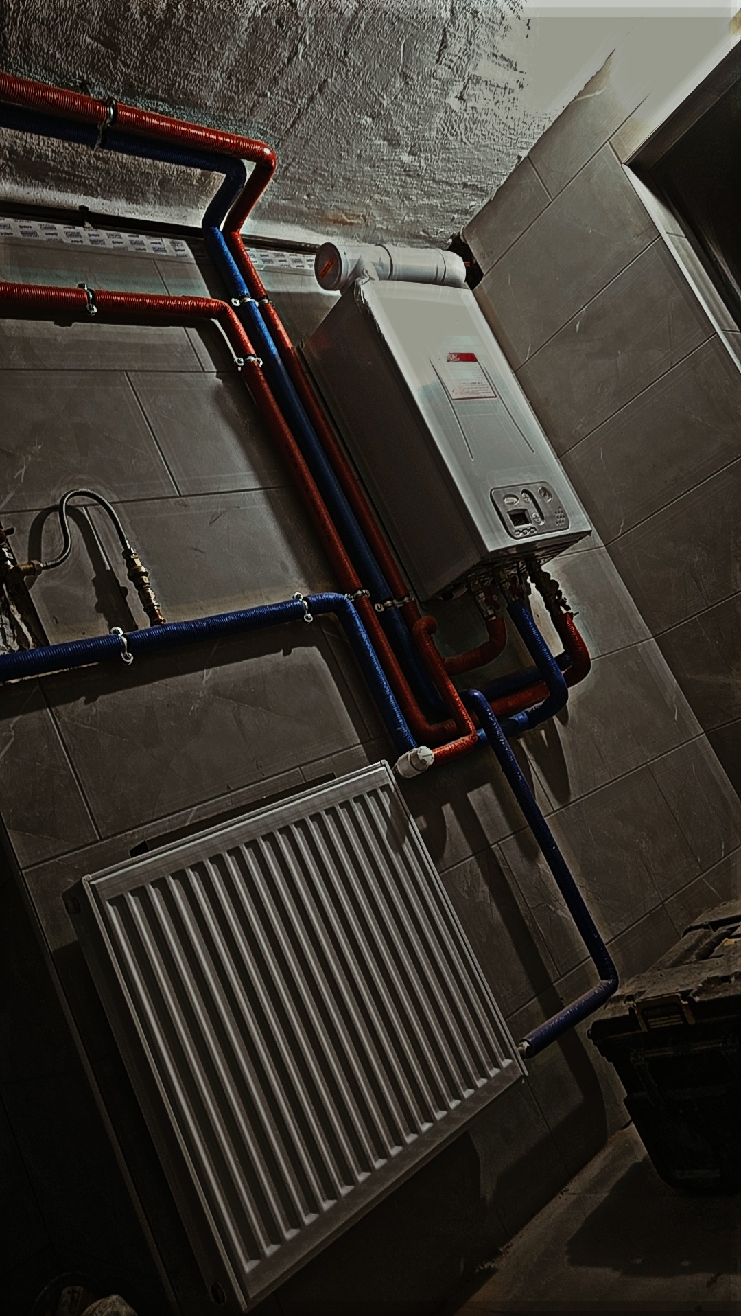 Zamontowany piec CO z podłączonymi rurami miedzianymi i plastikowymi, grzejnik panelowy na szarej płytce ściennej, widoczne elementy instalacji w pomieszczeniu.