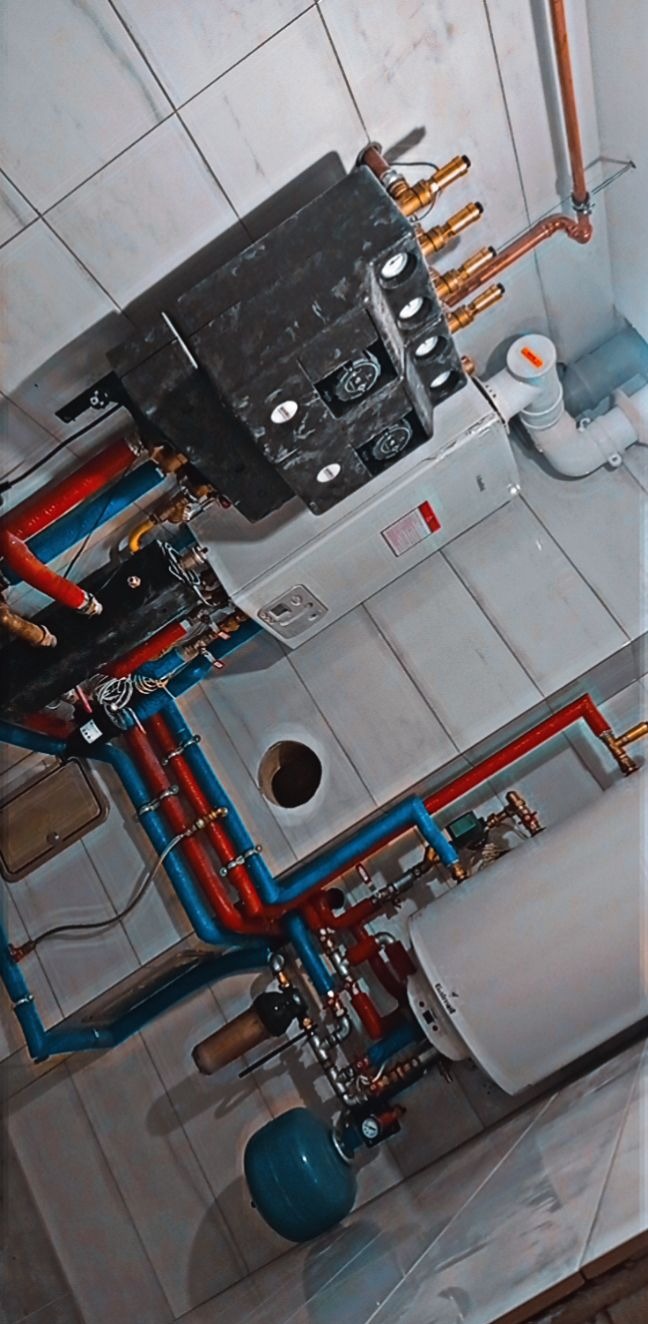 Kompleksowa instalacja kotła grzewczego z licznymi rurami w kolorach niebieskim i czerwonym, widok z dołu na tle białych płytek ceramicznych.