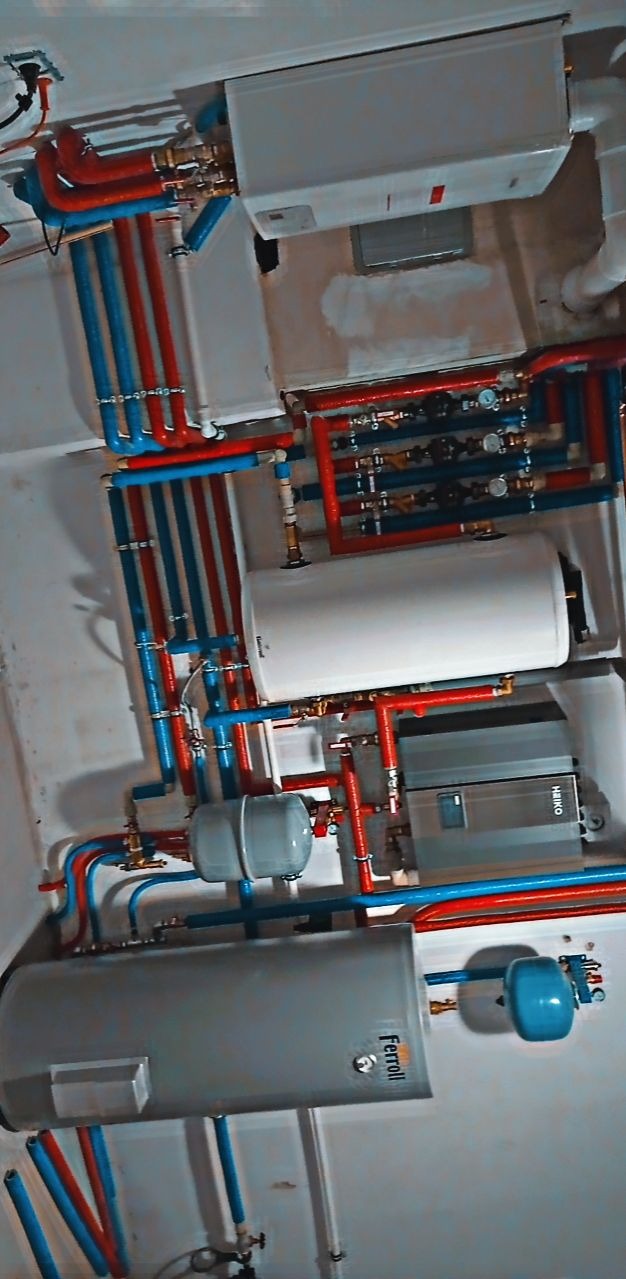 Skomplikowany układ instalacji grzewczej z rurami w kolorach czerwonym i niebieskim, zbiornikami, bojlerem Ferroli i piecem Haiko, widok z dołu.