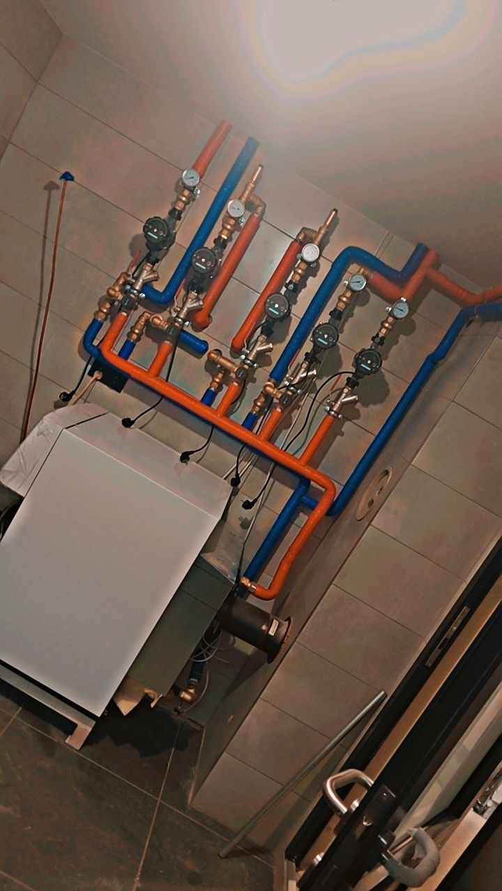 Nowoczesna instalacja grzewcza z piecem, rurami w kolorze pomarańczowym i niebieskim oraz manometrami, zamontowana na tle płytek ceramicznych.