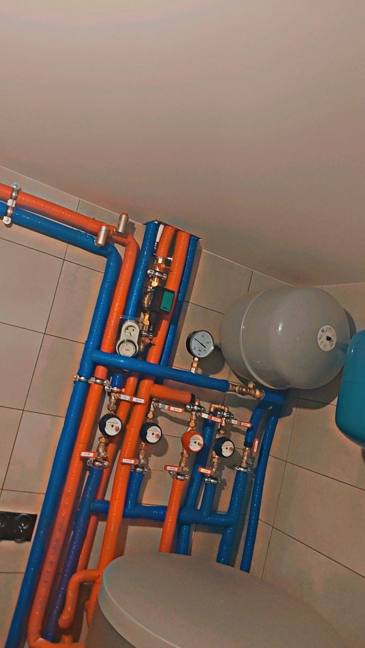 Rozbudowana instalacja wodna z rurami w izolacji termicznej w kolorach niebieskim i pomarańczowym, liczniki i manometry zamontowane na ścianie wyłożonej płytkami ceramicznymi, widoczny zbiornik...