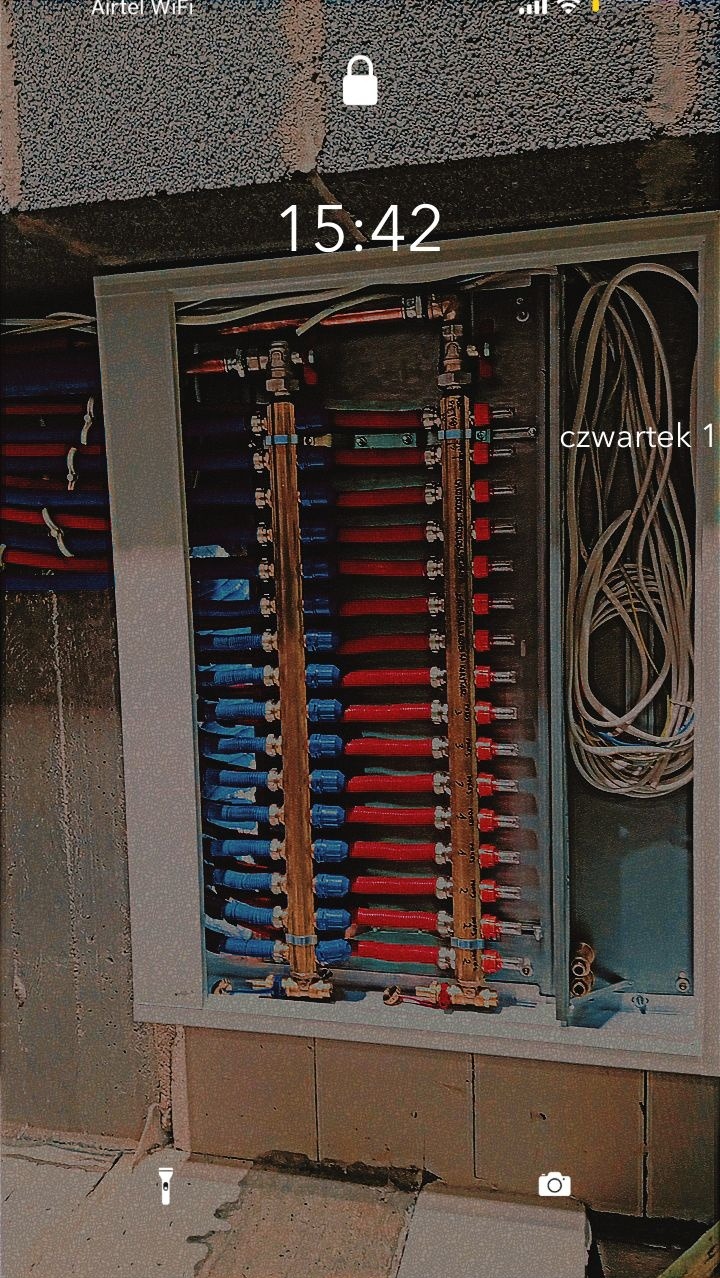 Rozdzielacz ogrzewania podłogowego z licznymi czerwonymi i niebieskimi rurkami, zamontowany w skrzynce w surowym stanie, widok z dołu na ścianę z widoczną fakturą betonu.