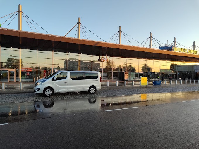 Biały bus Opel Vivaro zaparkowany przed przeszklonym budynkiem lotniska Katowice Airport z odbiciem w mokrej nawierzchni.