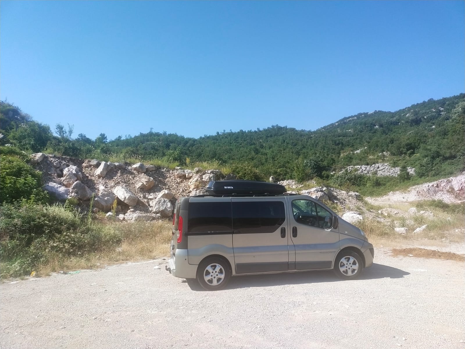 Szary van z czarnym boxem dachowym marki Taurus, zaparkowany na żwirowym placu w pobliżu skalistej skarpy porośniętej zielenią, błękitne niebo w tle.