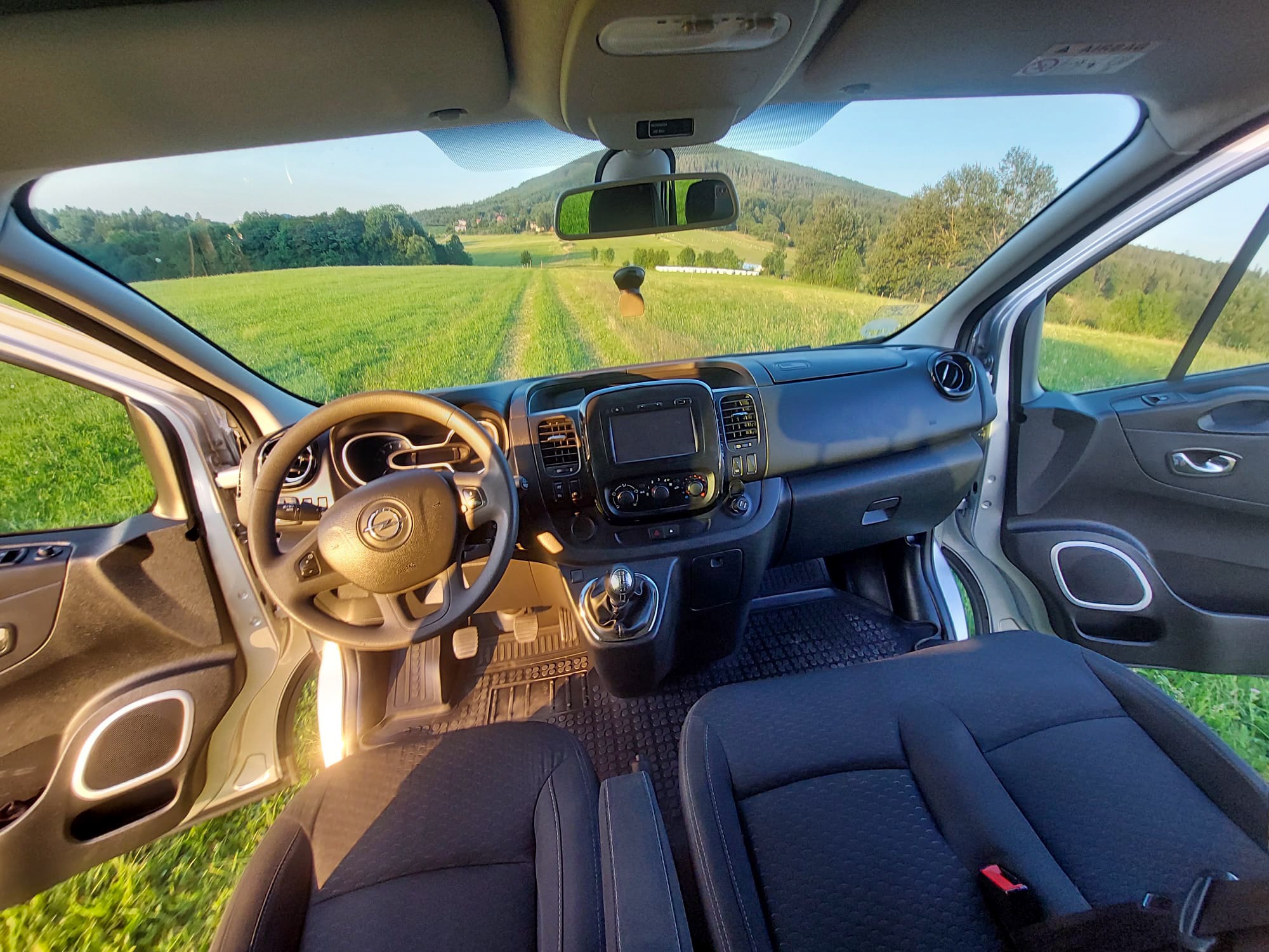 Wnętrze busa z widokiem na zielone pole i wzgórze, kierownica po lewej, otwarte drzwi po obu stronach.