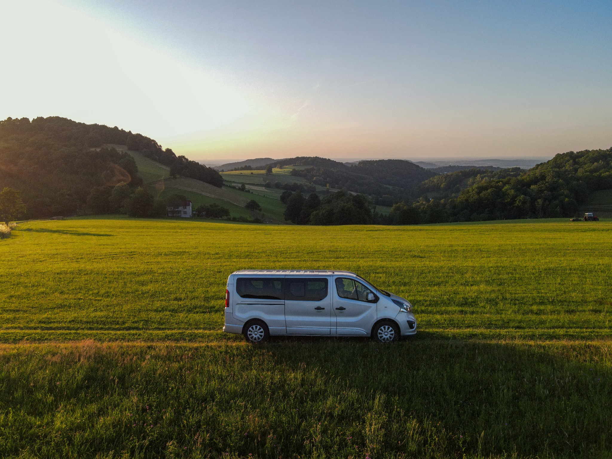 Srebrny bus na tle zielonego pola i pagórkowatego krajobrazu o zachodzie słońca, widok z lotu ptaka.