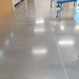 Wypolerowana, szara posadzka przemysłowa z widocznymi liniami podziału, odbijająca światło sufitowych lamp, z elementami konstrukcji w tle.