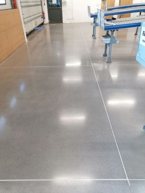 Wypolerowana, szara posadzka przemysłowa z widocznymi liniami podziału, odbijająca światło sufitowych lamp, z elementami konstrukcji w tle.