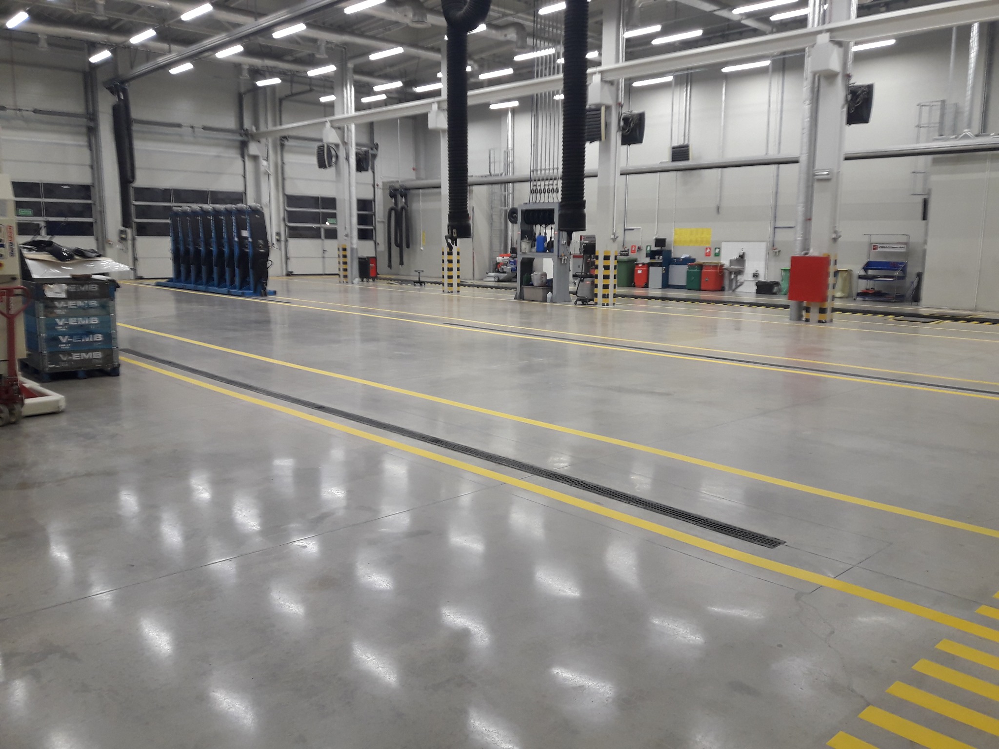 Gładka, szara posadzka przemysłowa z żółtymi liniami wyznaczającymi strefy, widoczne kratki odpływowe i wyposażenie hali produkcyjnej w tle.