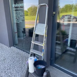 Czysty dom - Aluminiowa drabina przystawiona do szarej ściany obok okna z napisem 'drukujemy w małym i wielkim formacie', na drabince wiadro z wodą i pianą, ręcznik papierowy oraz butelka z płynem do mycia okien.