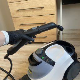 Czysty dom - Parownica Karcher w użyciu, dysza w dłoni w czarnej rękawicy, tło stanowią meble w jasnym odcieniu drewna.