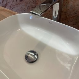 Czysty dom - Nowoczesna, biała umywalka nablatowa z chromowaną baterią marki Baliv, ustawiona na drewnianym blacie, na tle ściany wykończonej marmurem w odcieniach brązu i beżu.