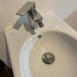 Biała umywalka Cersanit z chromowaną baterią bidetową o geometrycznym kształcie i widocznym korkiem spustowym.