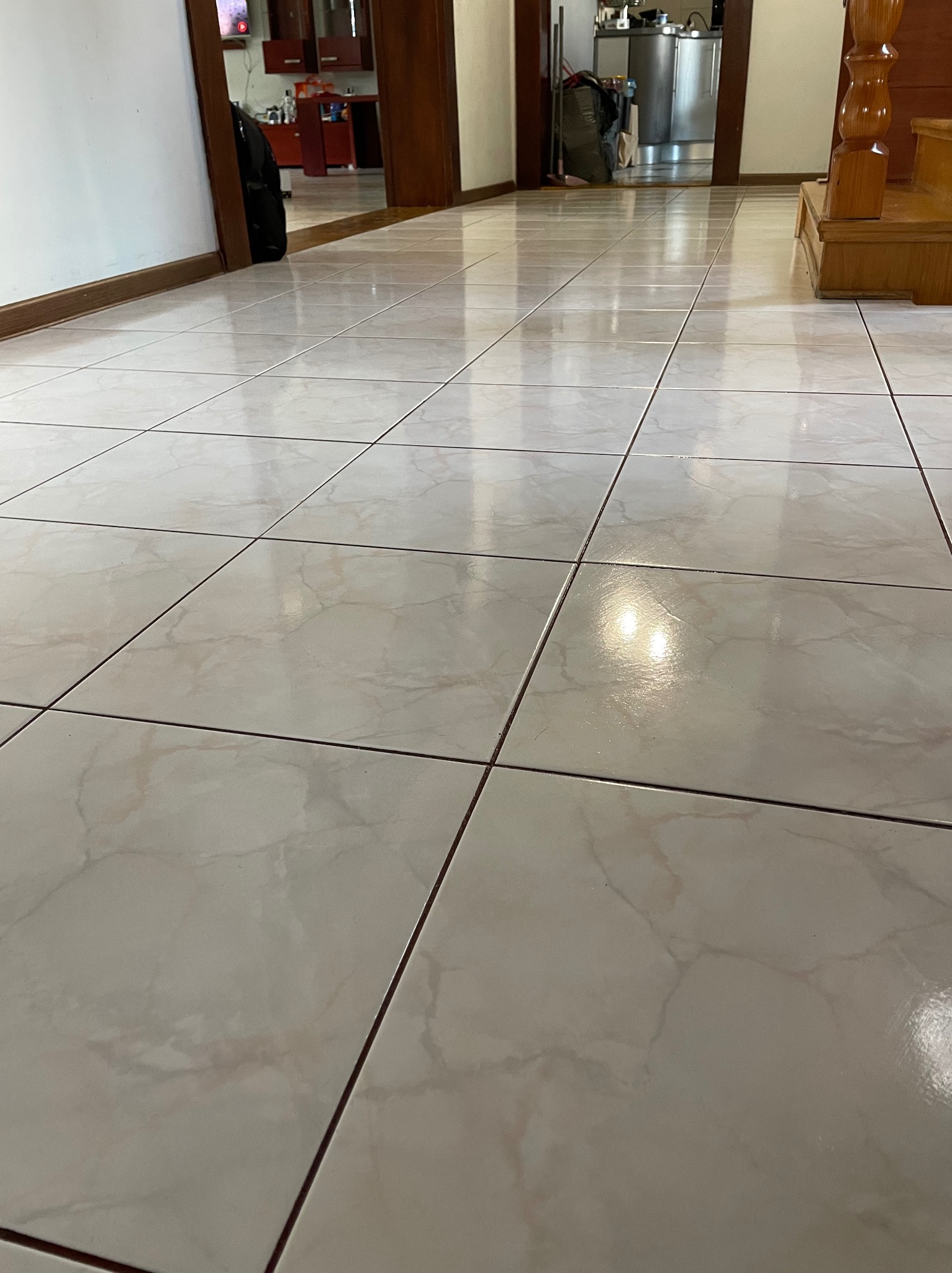 Błyszcząca, beżowa podłoga z płytek ceramicznych po umyciu, widoczna z niskiej perspektywy, z odblaskami światła i widocznymi fugami; w tle widoczne pomieszczenia domu.