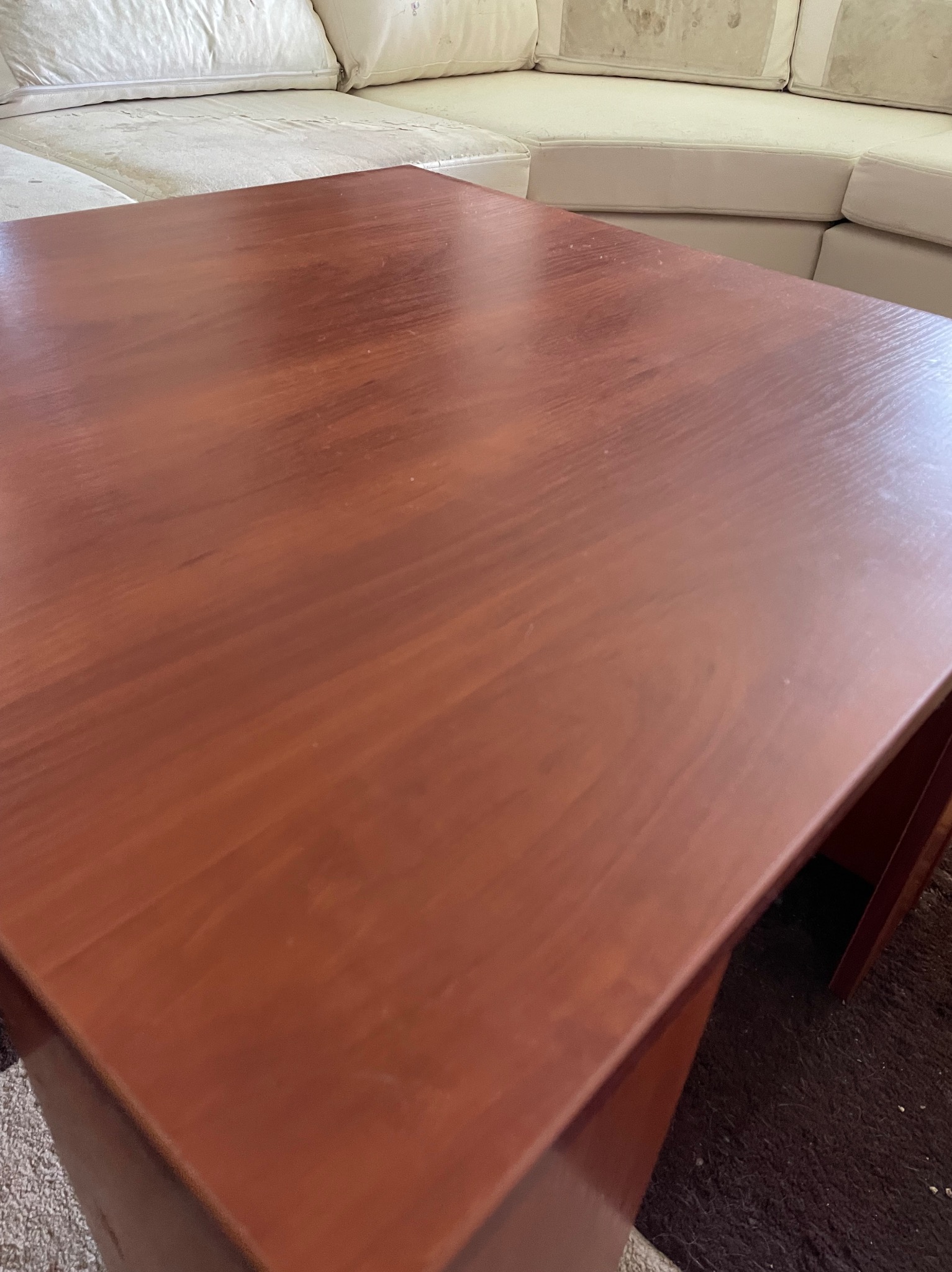 Ciemnobrązowy, drewniany stolik kawowy z widocznymi śladami użytkowania, stojący na ciemnym dywanie, z fragmentem brudnej, jasnej sofy w tle.