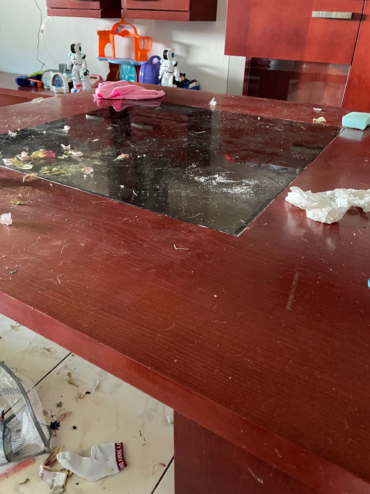 Brudny stół z ciemnym blatem, pokryty okruchami, płatkami kwiatów i drobnym kurzem. Na stole leżą również zmięte chusteczki i gąbka do czyszczenia. W tle widoczne zabawki i szafki. Na podłodze...