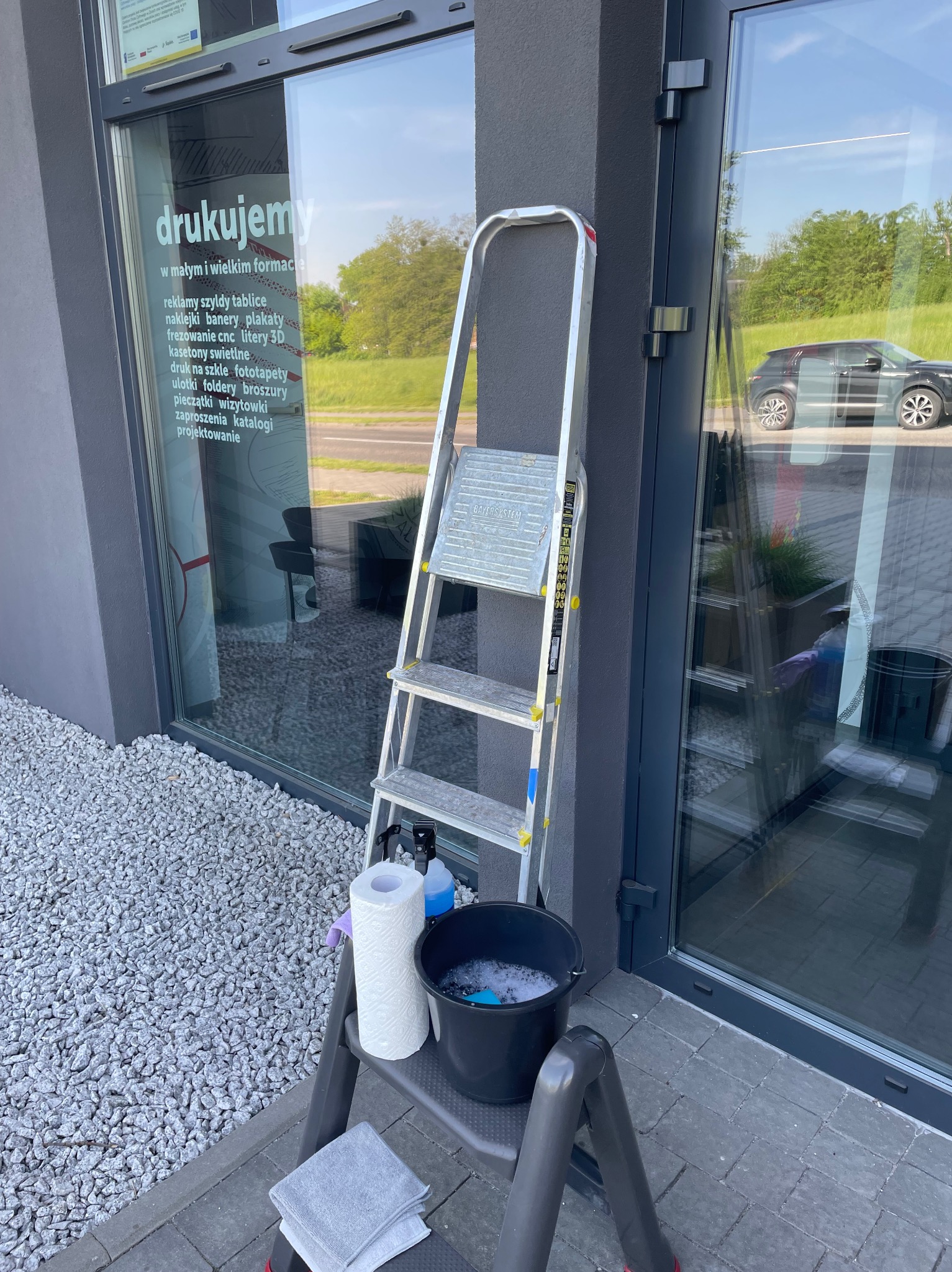 Aluminiowa drabina przystawiona do szarej ściany obok okna z napisem 'drukujemy w małym i wielkim formacie', na drabince wiadro z wodą i pianą, ręcznik papierowy oraz butelka z płynem do mycia okien.