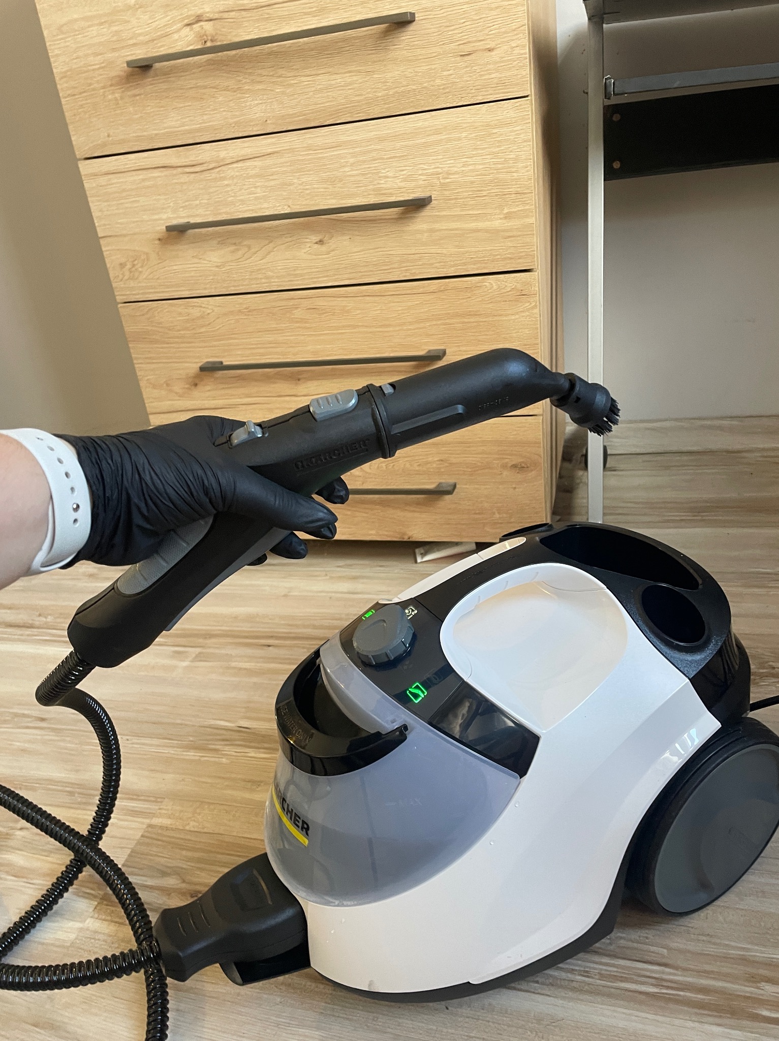 Parownica Karcher w użyciu, dysza w dłoni w czarnej rękawicy, tło stanowią meble w jasnym odcieniu drewna.