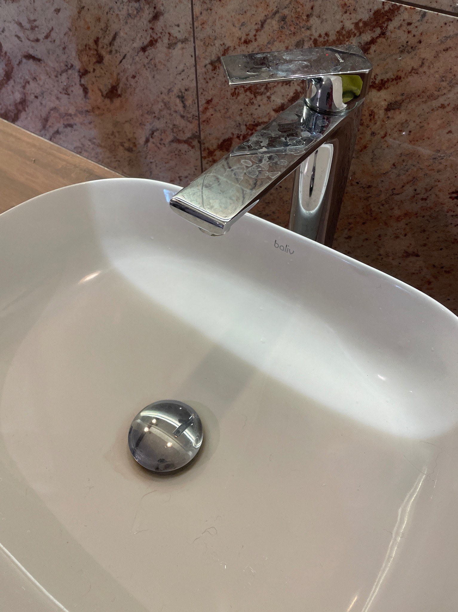 Biała, owalna umywalka z chromowaną baterią Baliv, na której widać ślady po wodzie. Marmurowa ściana w tle.