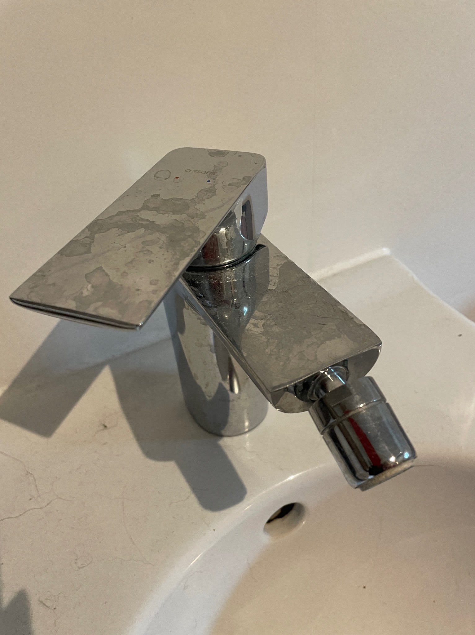Chromowana bateria umywalkowa marki Cersanit, pokryta zaciekami z kamienia, zamontowana na białej umywalce.