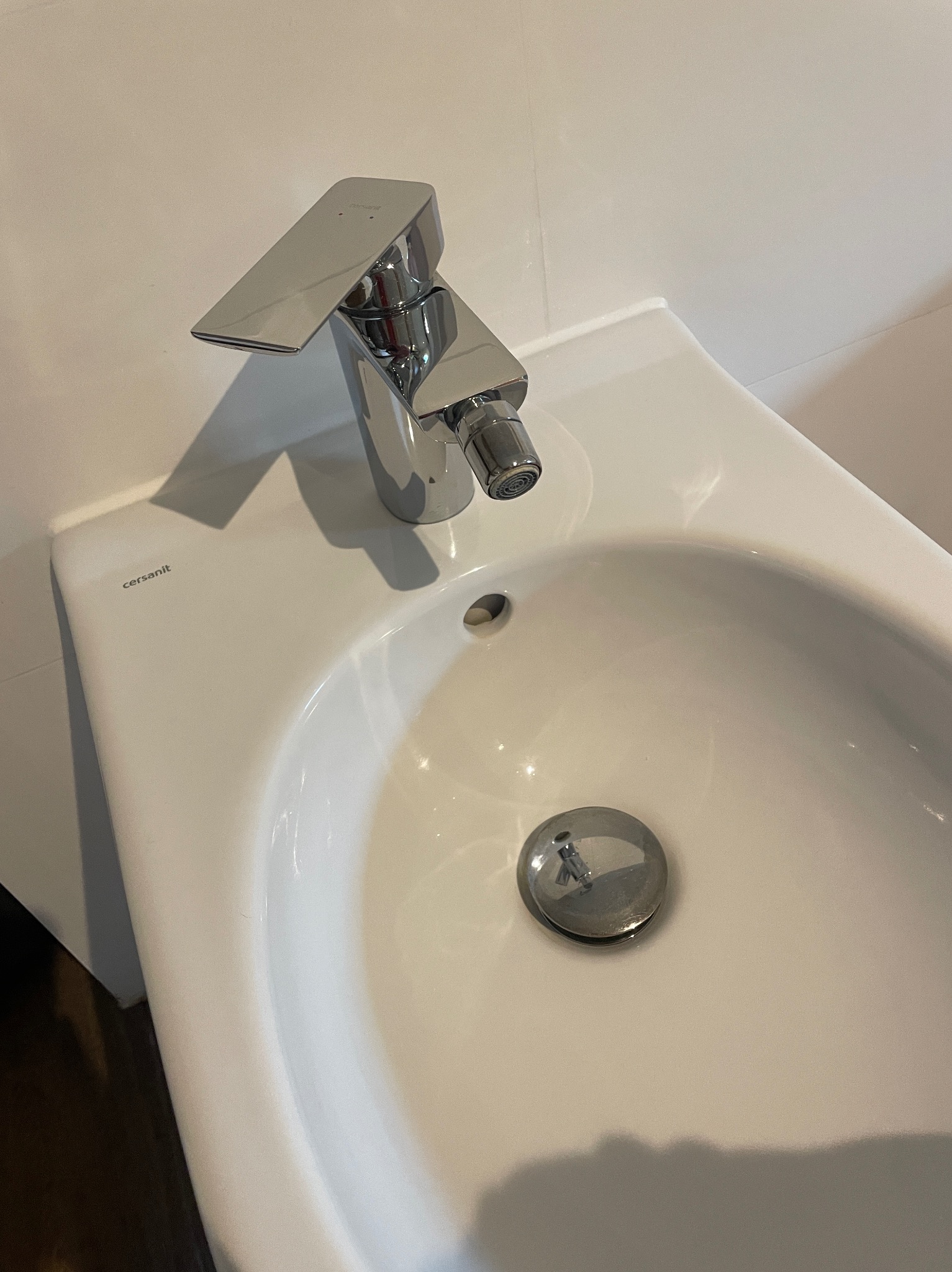 Biała umywalka Cersanit z chromowaną baterią bidetową o geometrycznym kształcie i widocznym korkiem spustowym.