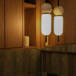 Wiktoria Surgota Moono Design - Nowoczesna łazienka z drewnianą szafką, podłużnymi płytkami w odcieniach brązu i dwoma wiszącymi lampami z kulistymi elementami. Minimalistyczny design.