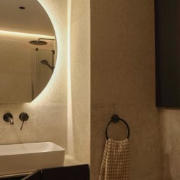 Wiktoria Surgota Moono Design - Nowoczesna łazienka z okrągłym lustrem LED, minimalistyczną umywalką i beżowymi płytkami. W tle widoczny prysznic. Ciepłe oświetlenie tworzy przytulny klimat.