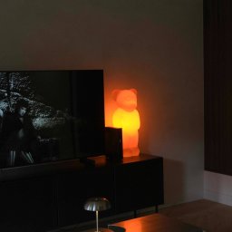 Wiktoria Surgota Moono Design - Nowoczesny salon z czarnymi meblami, telewizorem i lampą w kształcie misia, oświetlającą wnętrze ciepłym, pomarańczowym światłem. Metalowa lampka na stoliku kawowym.