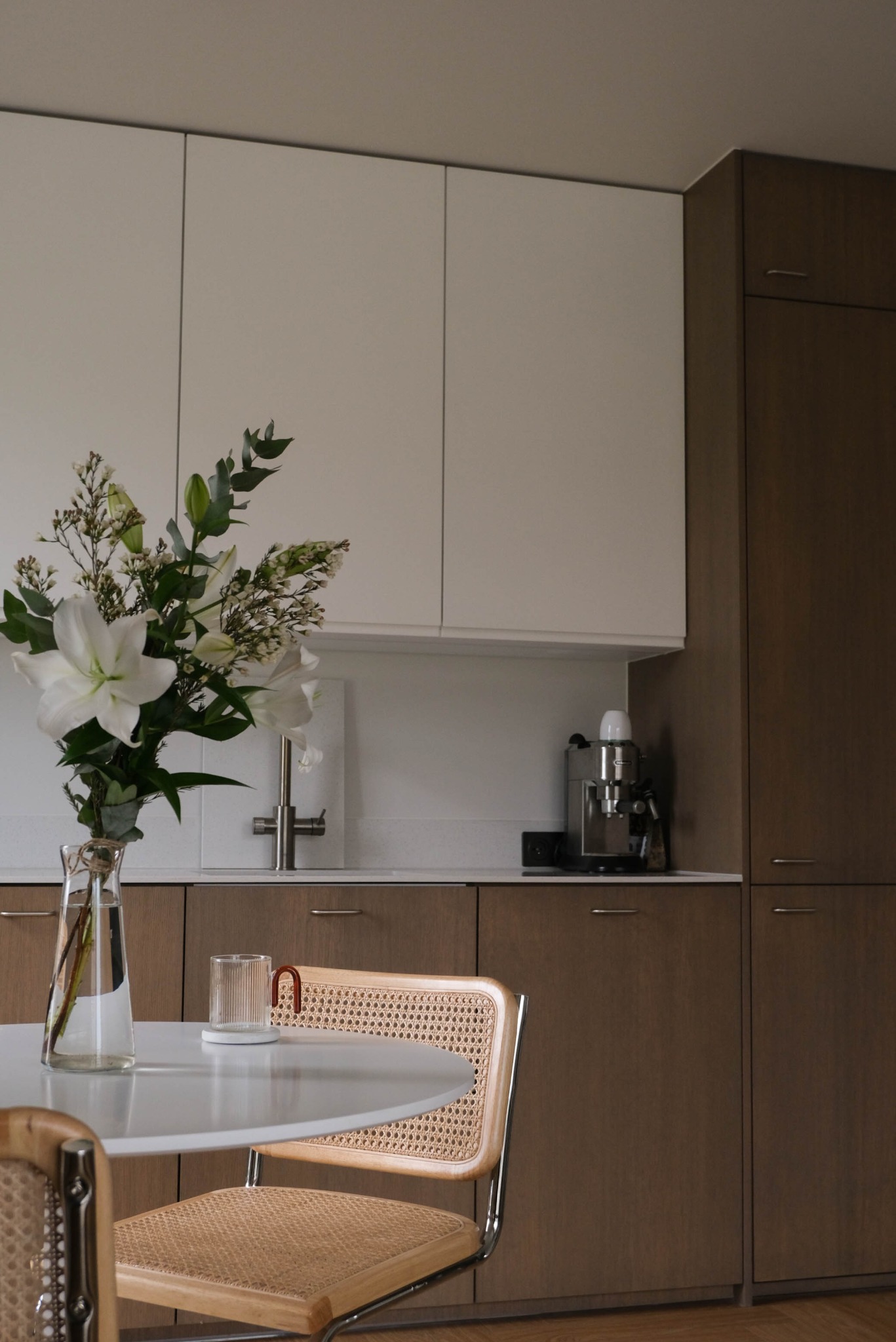 Minimalistyczna kuchnia z białymi i drewnianymi szafkami, okrągłym stołem z bukietem lilii i krzesłami z rattanu. Ekspres do kawy na blacie.