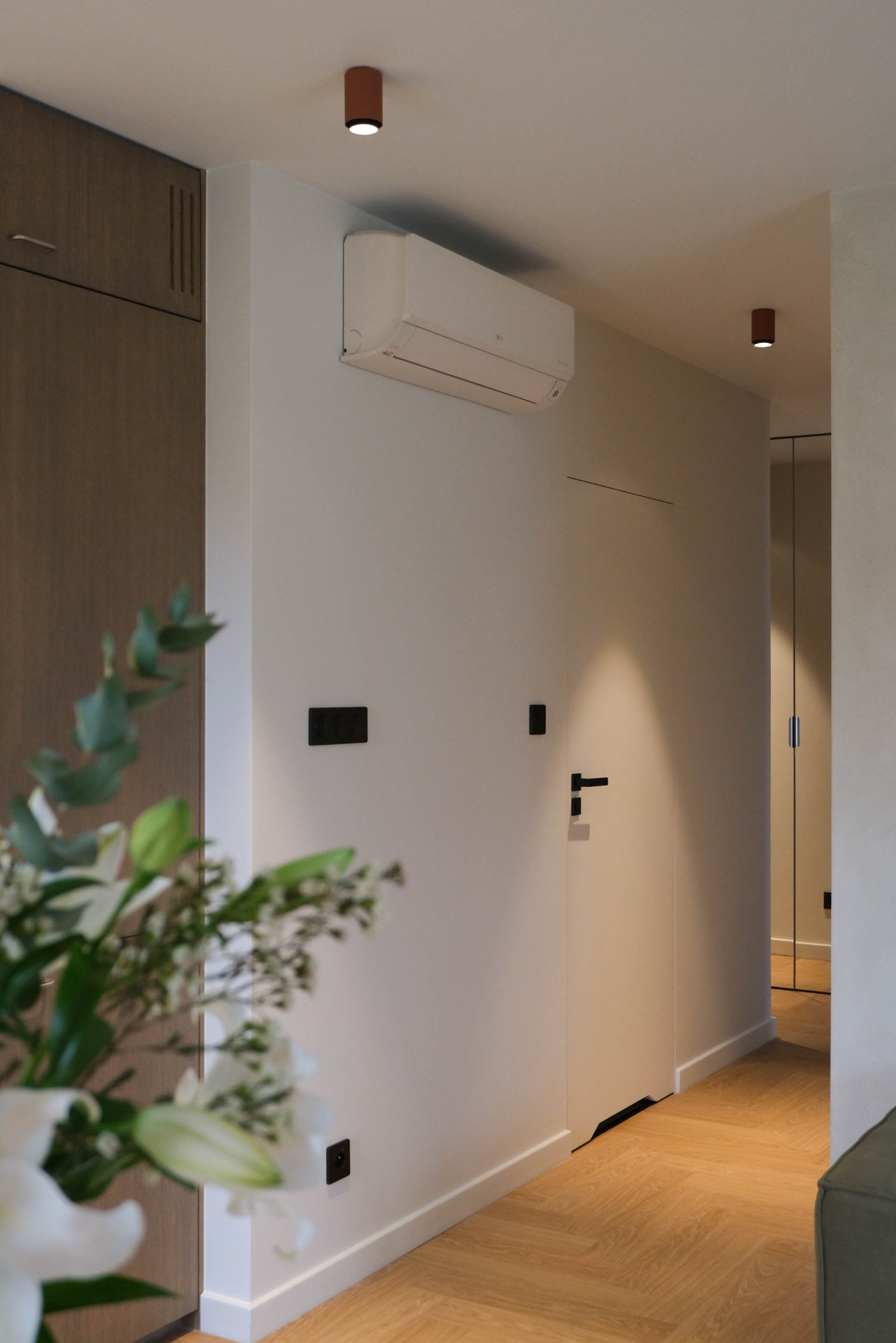 Minimalistyczny przedpokój: parkiet w jodełkę, białe ściany z ukrytymi drzwiami, czarne akcenty (gniazdka, włączniki, klamka) i klimatyzator. W tle lustrzane drzwi.