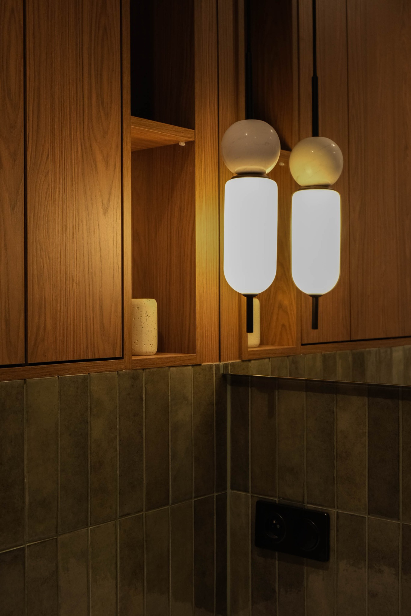 Nowoczesna łazienka z drewnianą szafką, podłużnymi płytkami w odcieniach brązu i dwoma wiszącymi lampami z kulistymi elementami. Minimalistyczny design.