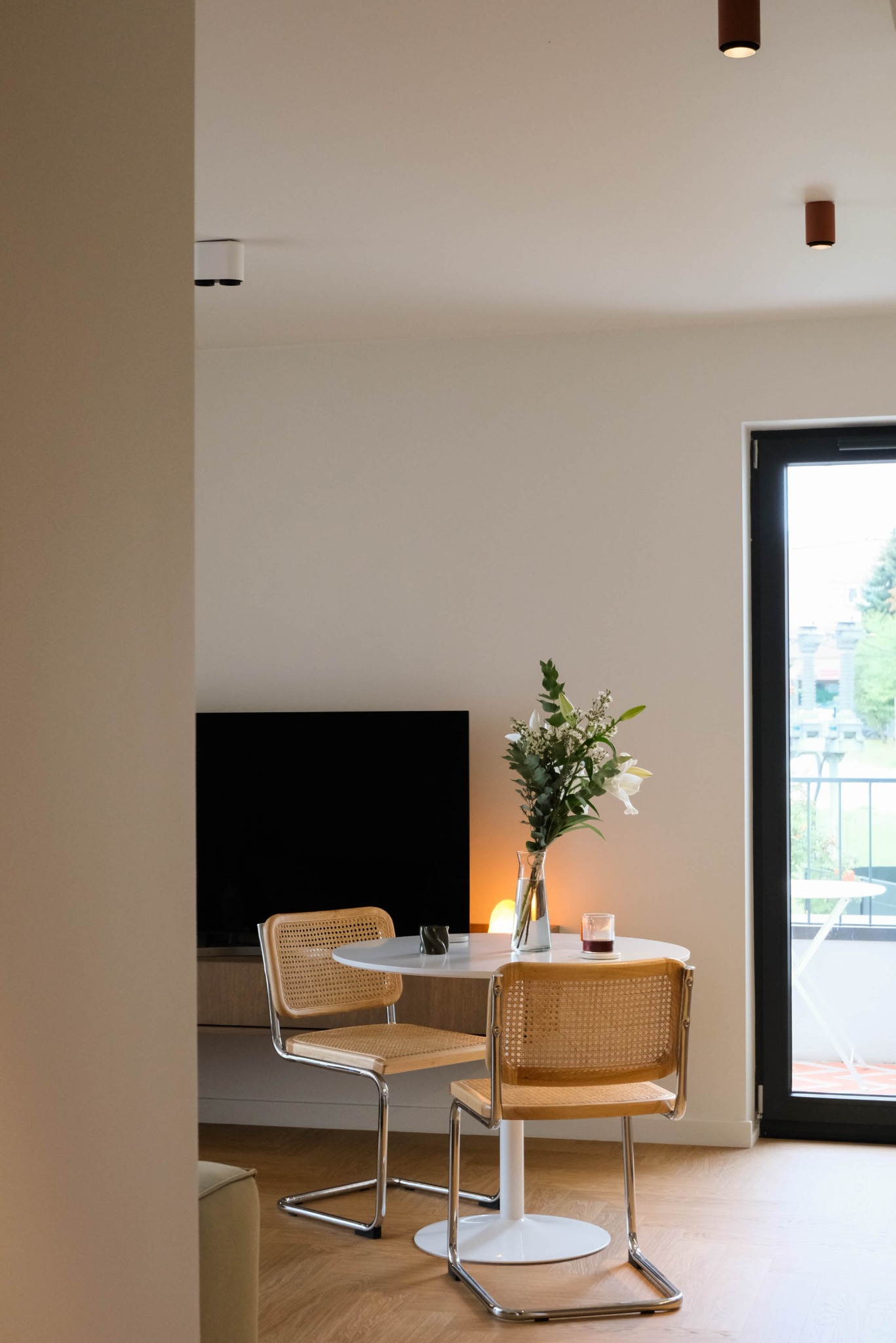 Jasne wnętrze mieszkania z okrągłym stołem, krzesłami z rattanu i bukietem kwiatów. Minimalistyczny design, parkiet na podłodze, widok na balkon przez okno.