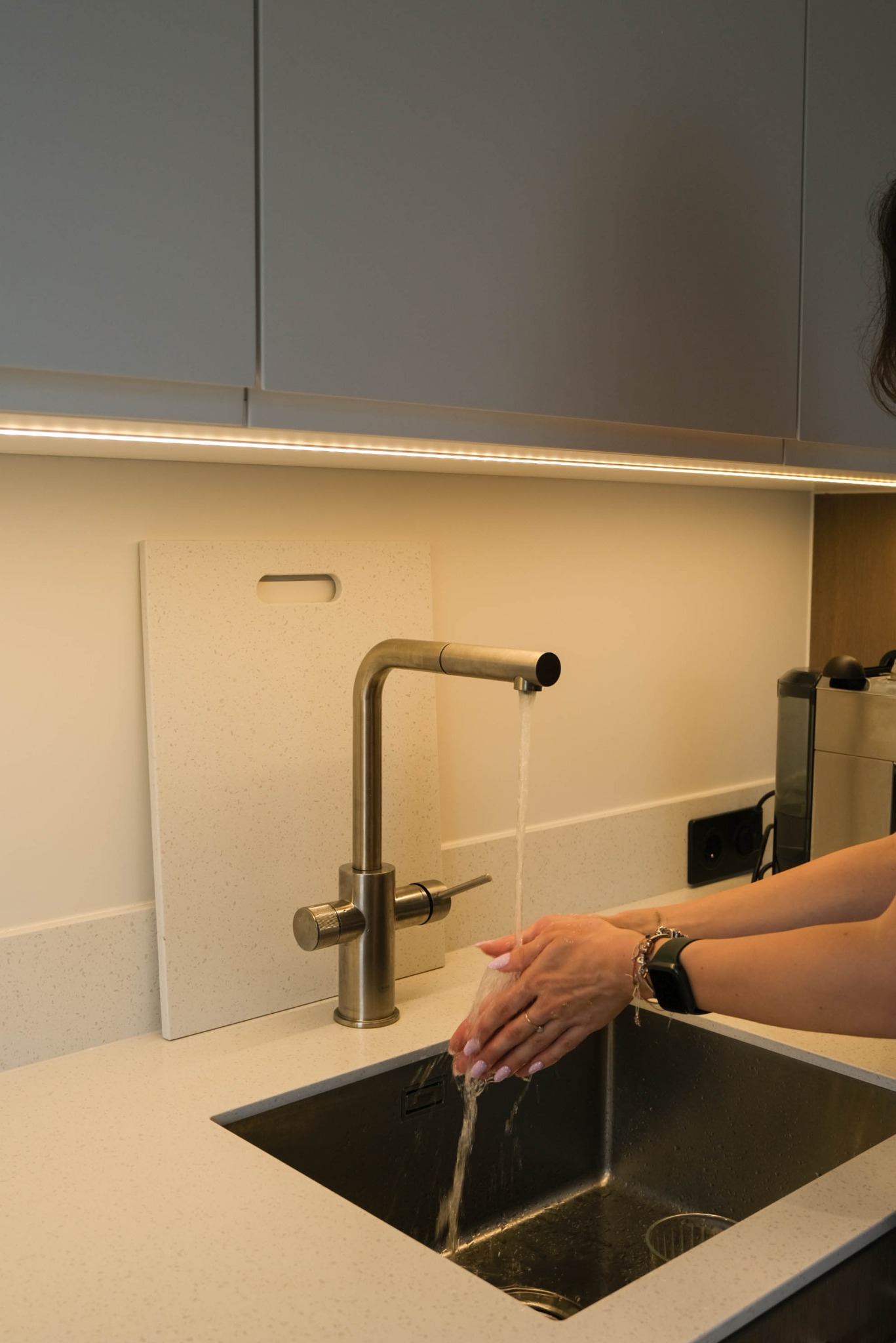 Kuchenny zlew z baterią w odcieniach szarości, woda płynie na dłonie kobiety z zegarkiem. W tle podświetlane szafki i ekspres do kawy.