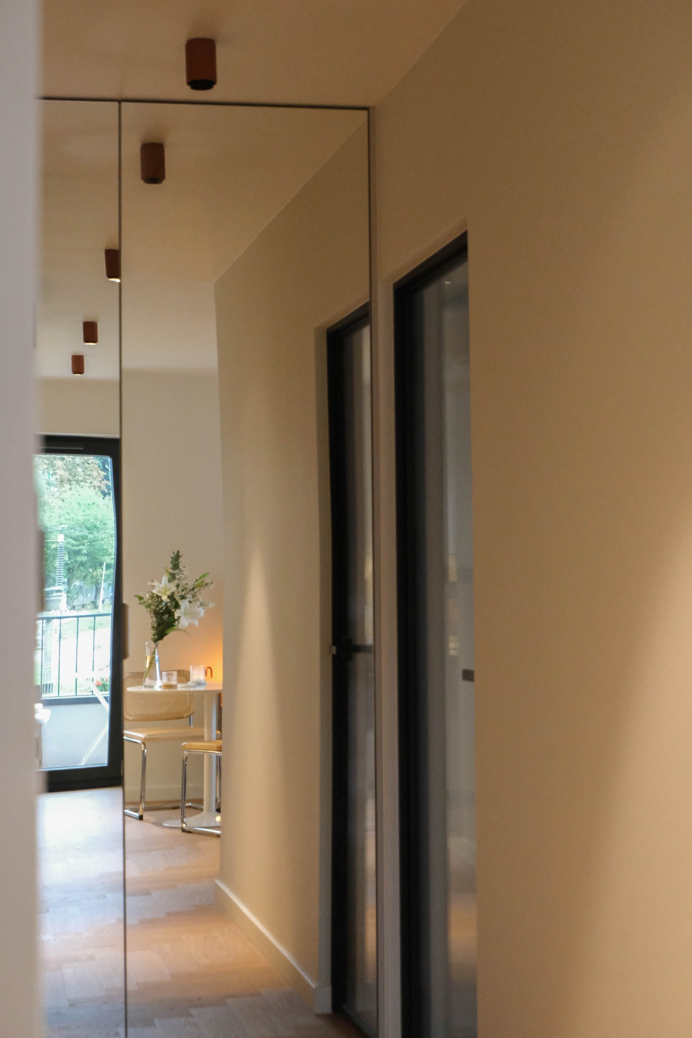 Lustro w przedpokoju odbija jasne wnętrze mieszkania z drewnianą podłogą, stolikiem i kwiatami. Widoczne drzwi z czarną ramą i oświetlenie sufitowe.