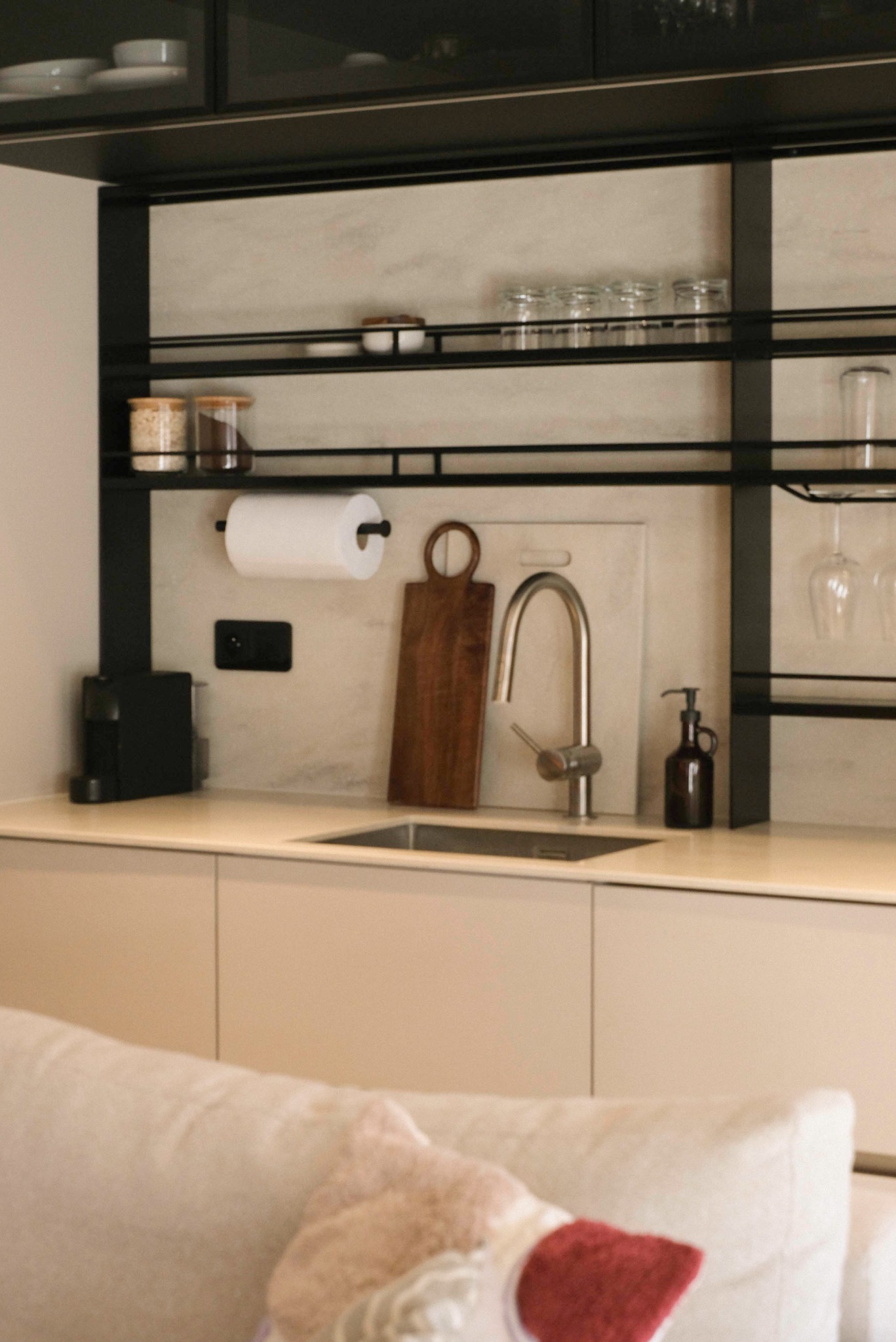 Minimalistyczna kuchnia z beżowymi szafkami, czarnymi metalowymi półkami i drewnianymi akcentami. Widok z salonu, fragment sofy z poduszką.