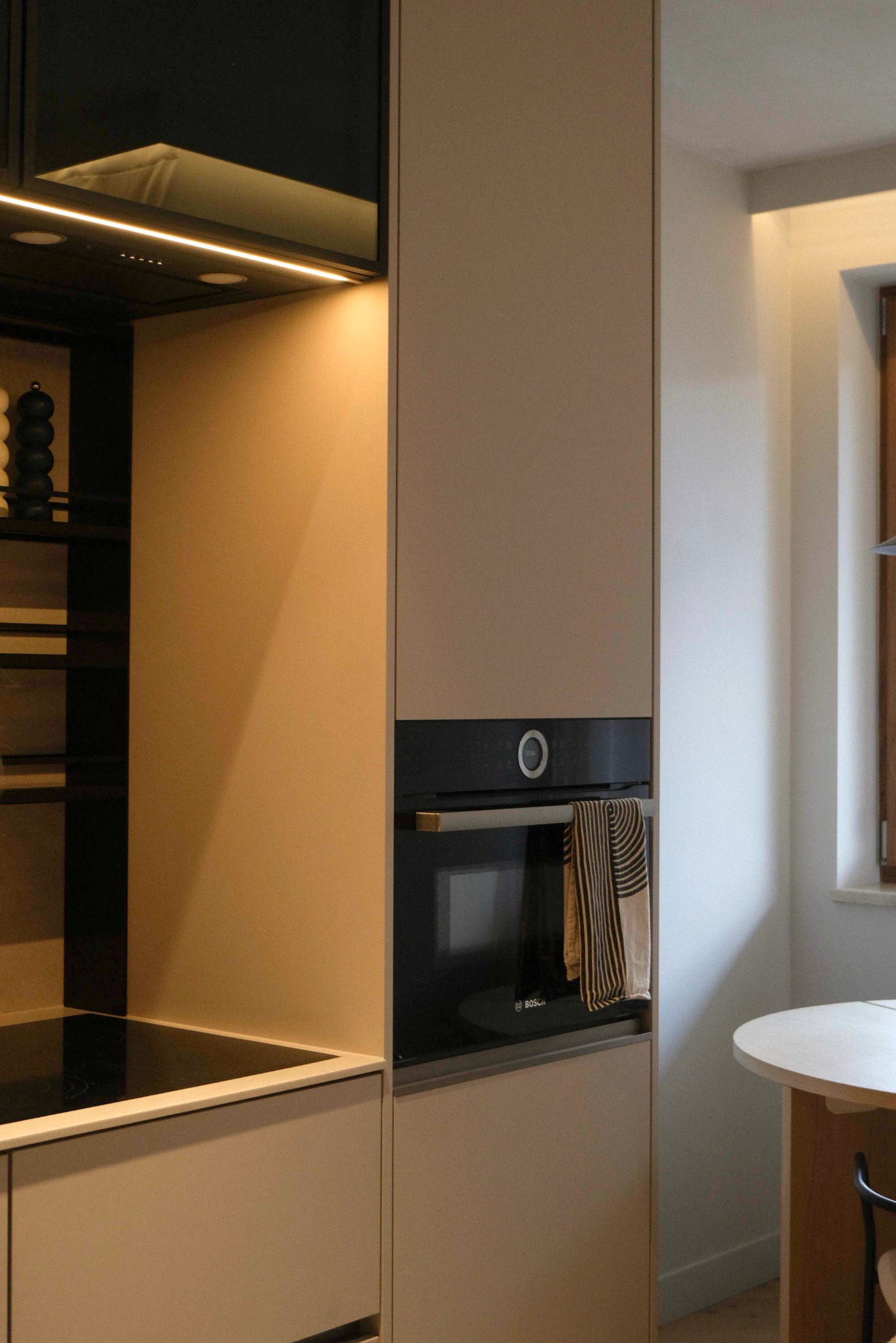 Minimalistyczna zabudowa kuchenna w beżowej tonacji z czarnym piekarnikiem i płytą grzewczą. Oświetlenie LED podkreśla nowoczesny charakter wnętrza.