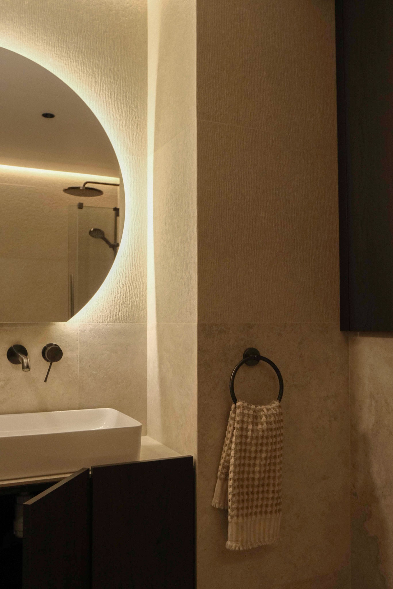 Nowoczesna łazienka z okrągłym lustrem LED, minimalistyczną umywalką i beżowymi płytkami. W tle widoczny prysznic. Ciepłe oświetlenie tworzy przytulny klimat.